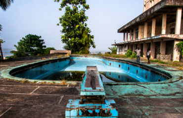 Piscina vacía y deteriorada rodeada de vegetación, frente a un edificio abandonado.