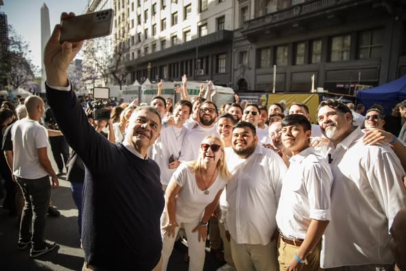 Grupo de personas sonriendo y posando para una selfie en un evento al aire libre, con edificios de fondo y el Obelisco de Buenos Aires a la distancia.