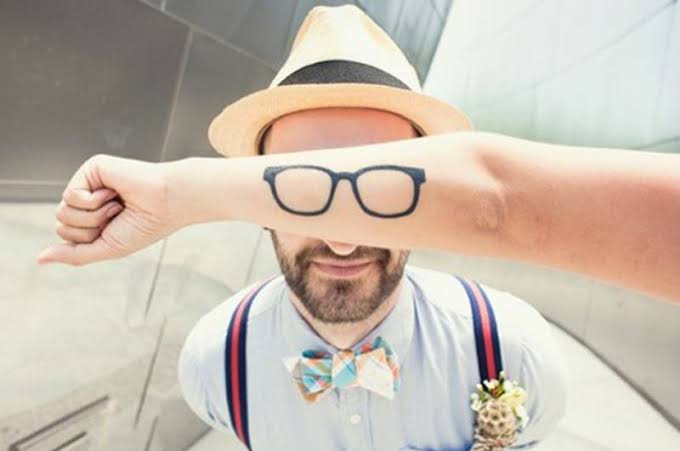Hombre con barba y sombrero en primer plano, con un brazo que sostiene una mano mostrando un tatuaje de gafas en el antebrazo.