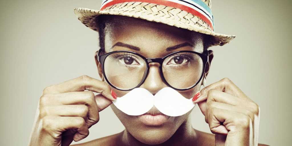Persona con gafas y sombrero sosteniendo un bigote de papel frente a su cara.