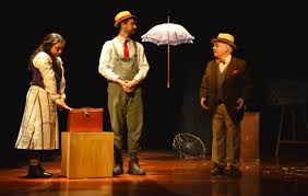 Tres actores en un escenario, uno de pie junto a una caja, otro con un paraguas colgante y el tercero observando, todos vestidos con trajes vintage.