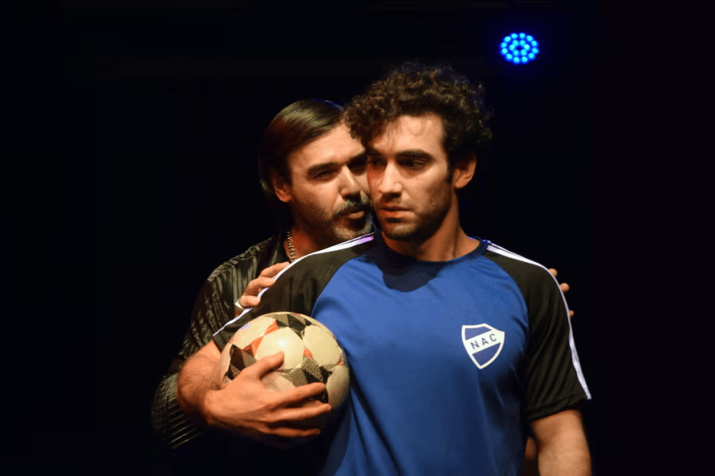 Dos hombres en un escenario, uno de pie sosteniendo un balón de fútbol y el otro detrás, sugiriendo una interacción intensa.