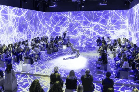 Performance en un espacio inmersivo con proyecciones de luz abstracta, rodeado de un público atento sentado en plataformas.