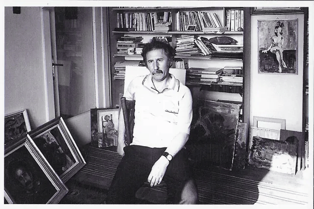 Hombre sentado en una habitación con estantes de libros y varias pinturas enmarcadas alrededor.