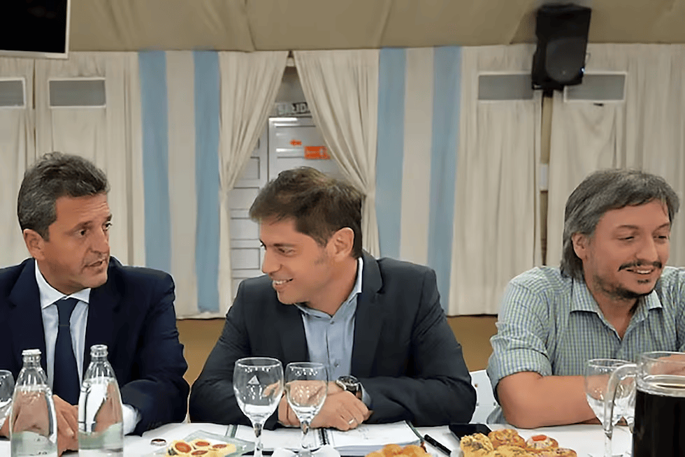 Tres hombres sentados en una mesa durante una reunión, con comida y bebida frente a ellos. Fondo con cortinas y decoraciones relacionadas con Argentina.