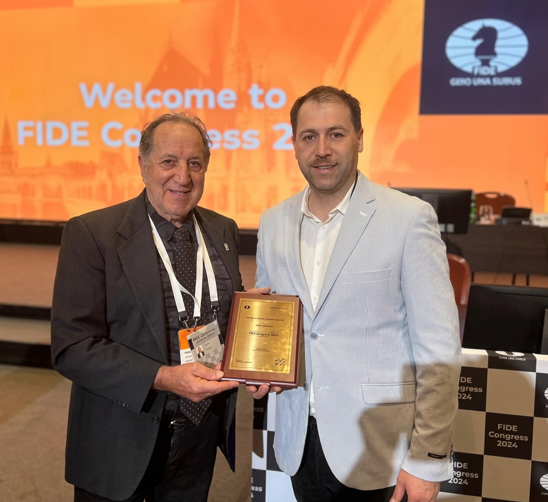 Dos hombres sonrientes sosteniendo un premio en el Congreso de la FIDE 2024, con un fondo que dice 'Welcome to FIDE Congress 2024'.