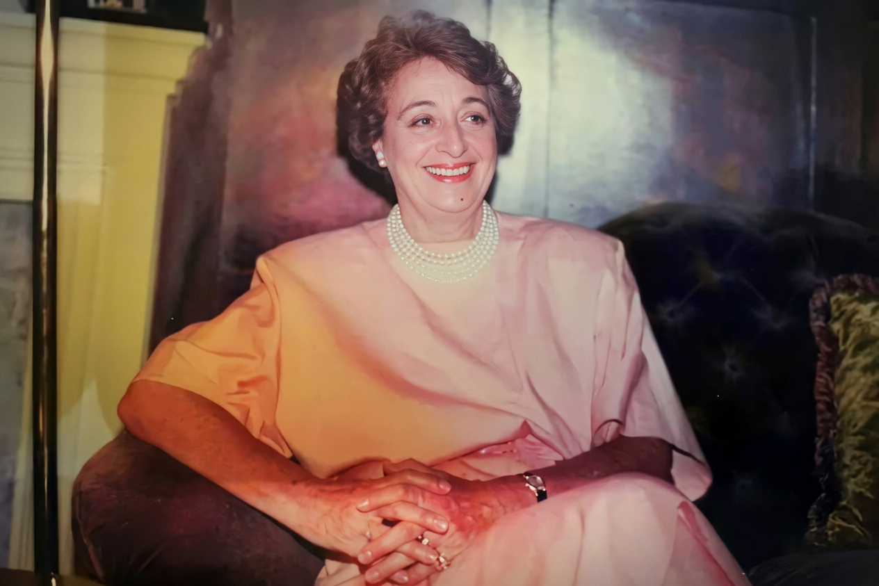 image Mujer sentada con una blusa rosa y collar de perlas, sonriendo, en un ambiente elegante.