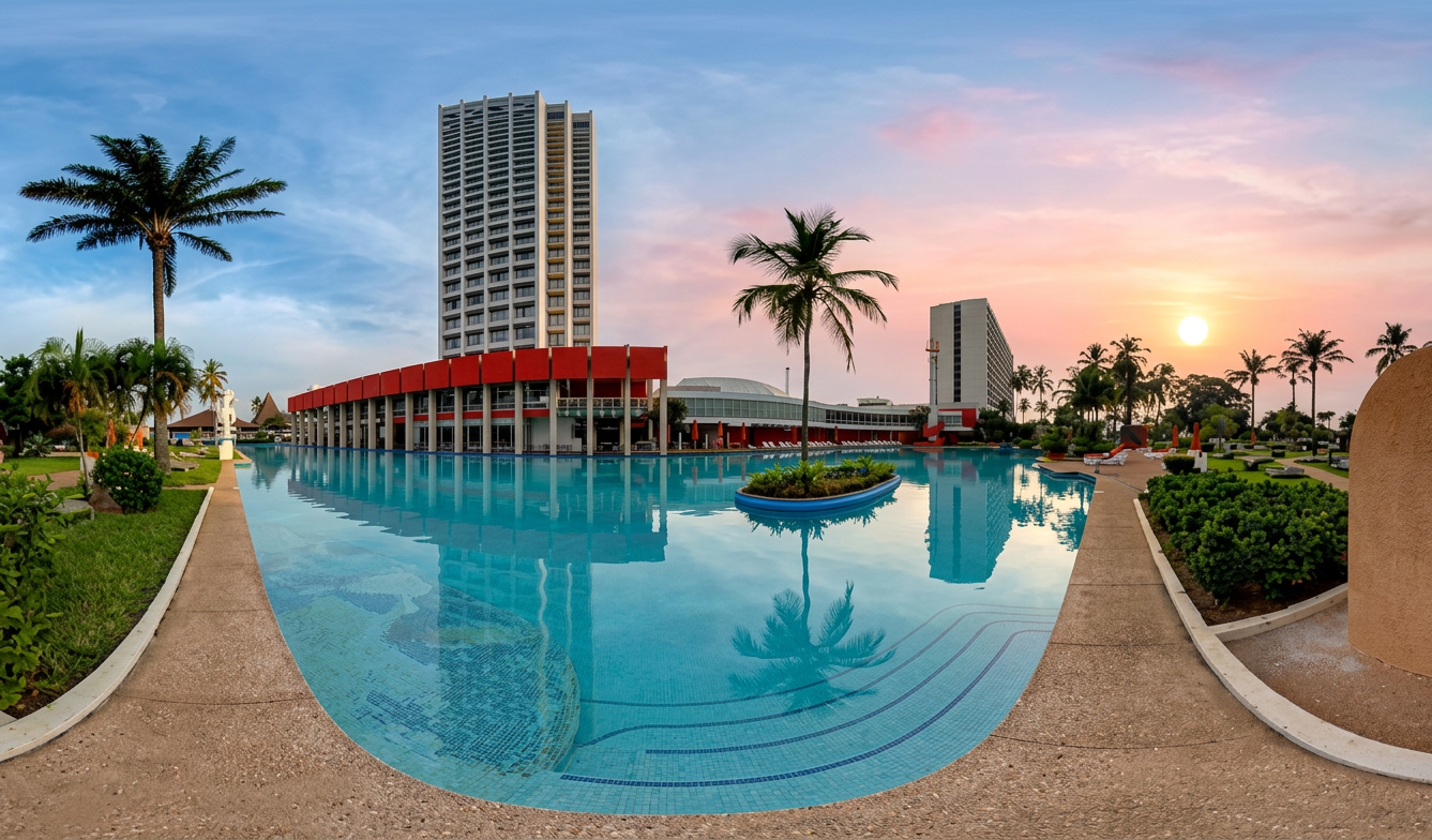 Vista panorámica de un hotel junto a una piscina, con palmeras y un atardecer colorido en el cielo.