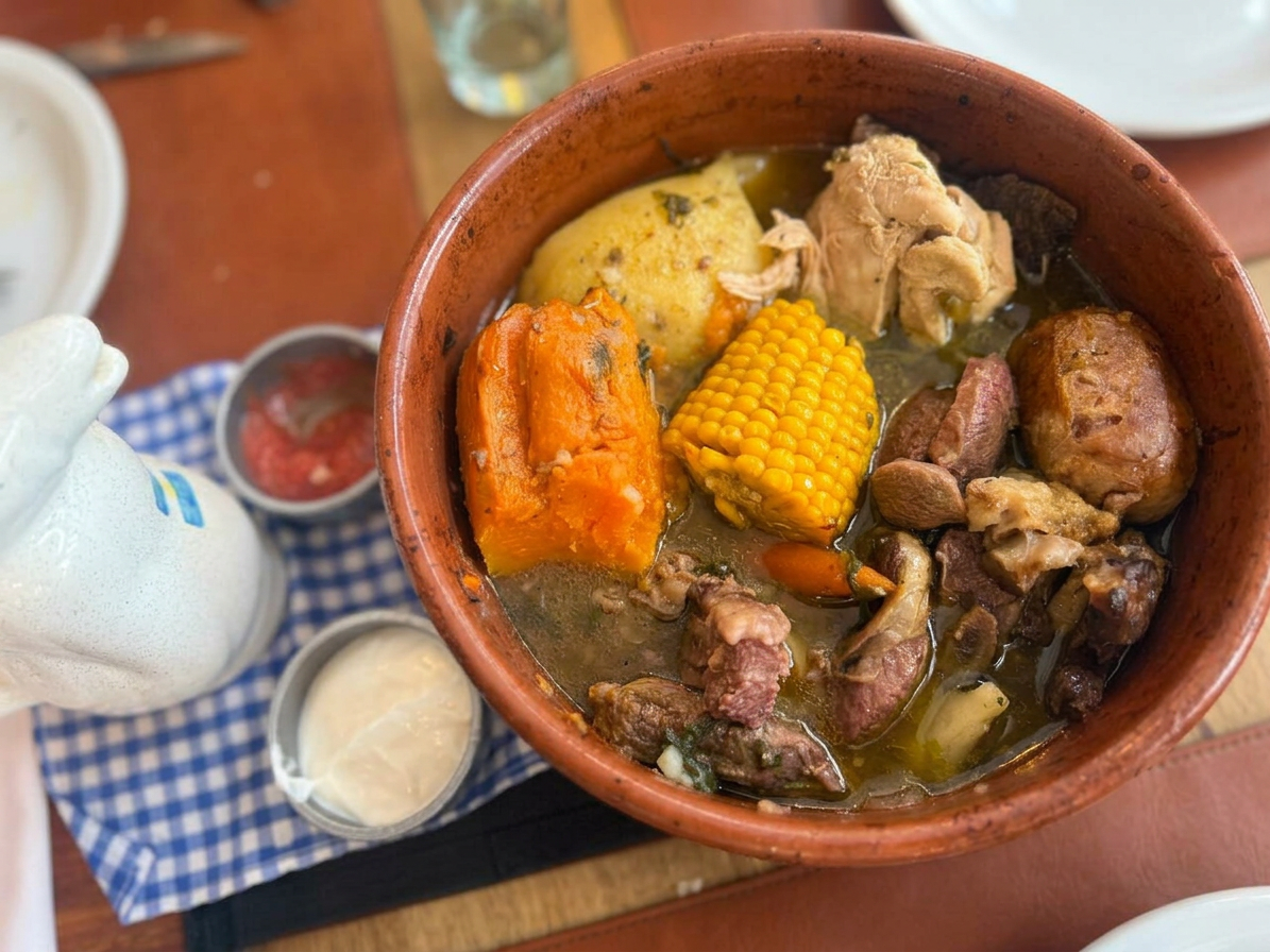 puchero Un plato de sopa tradicional servido en un tazón de barro, con carne, maíz, y verduras como zanahoria y calabaza.