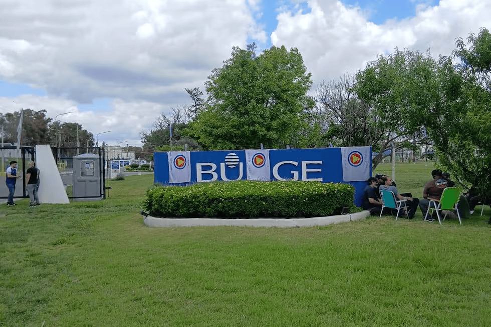 Vista de la entrada a un lugar con una señal que dice 'BÜGE', rodeada de un área verde y personas sentadas en sillas.