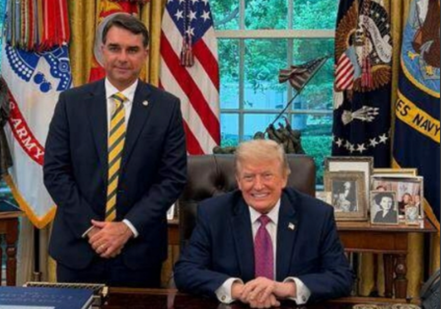 El expresidente Donald Trump sentado en un escritorio en la Oficina Oval, sonriendo, acompañado por un hombre de pie a su lado, con fondo de banderas y decoraciones presidenciales.