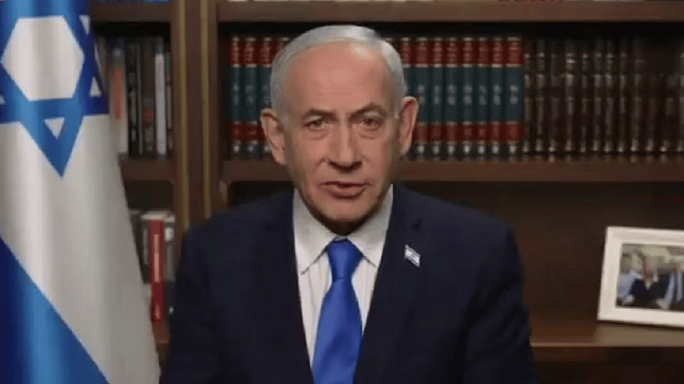 Hombre con traje oscuro y corbata azul, sentado frente a un fondo de estanterías con libros y una foto enmarcada, junto a una bandera de Israel.