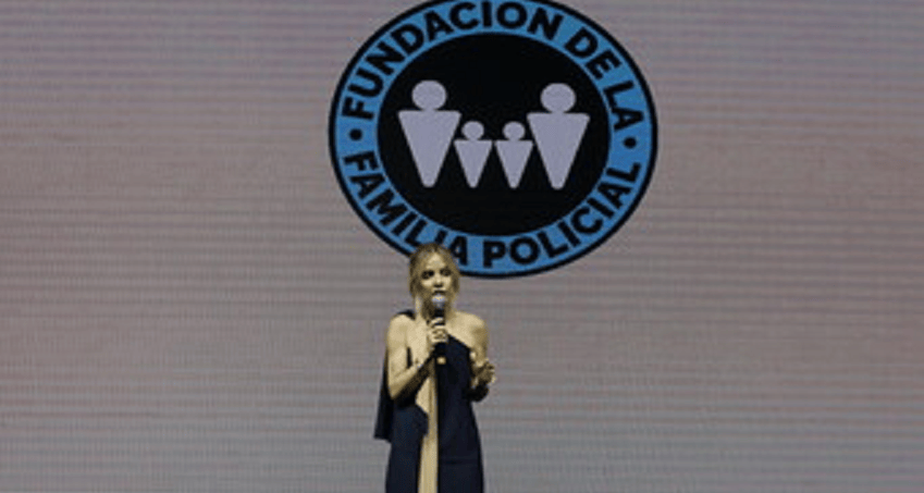 Una persona hablando en un micrófono frente a una pantalla con el logo de la Fundación de la Familia Policial.