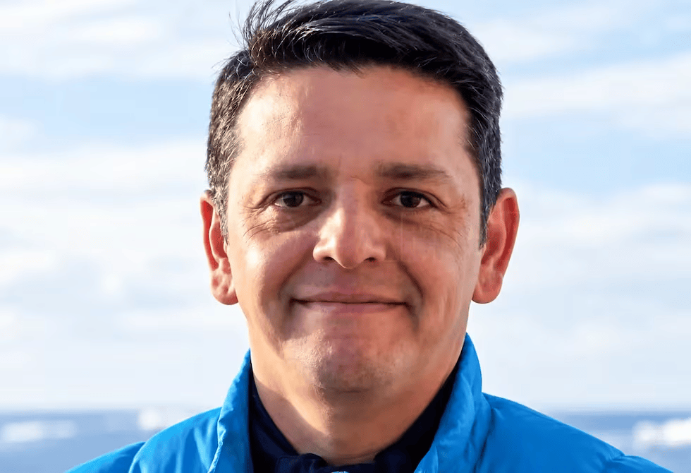 Hombre sonriendo con chaqueta azul, mar y cielo al fondo.