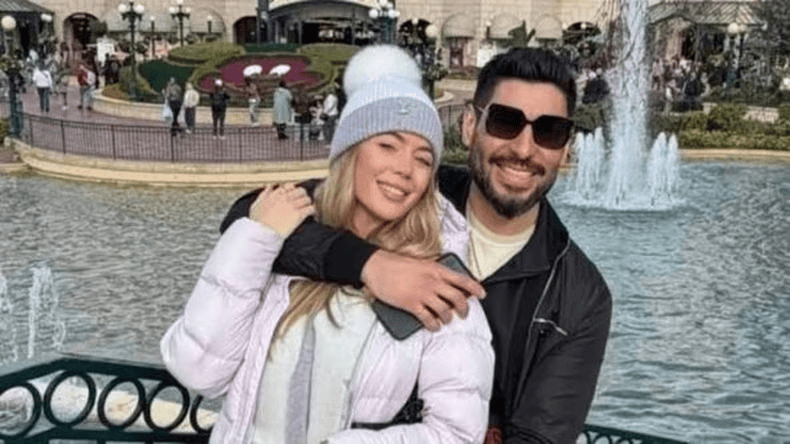 Una pareja sonriente posando frente a una fuente en un parque, rodeada de gente.