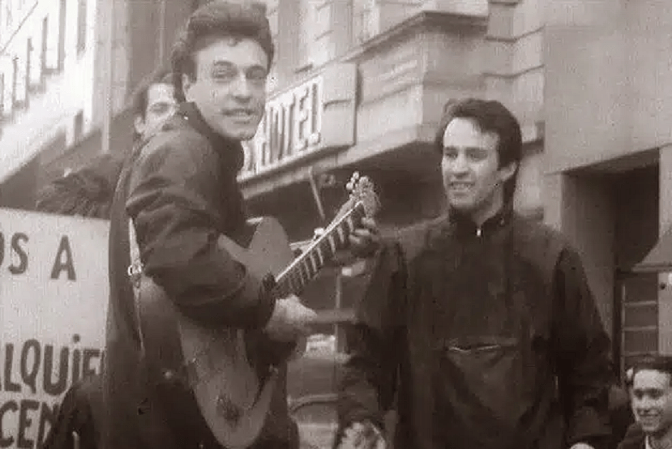 Dos hombres en una calle, uno tocando la guitarra y sonriendo, mientras el otro lo observa con alegría. Al fondo hay un cartel que dice '¡ALQUILER!'.