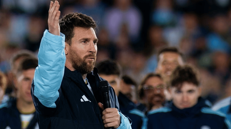Lionel Messi hablando con un micrófono, levantando la mano en un evento, rodeado de otros jugadores.