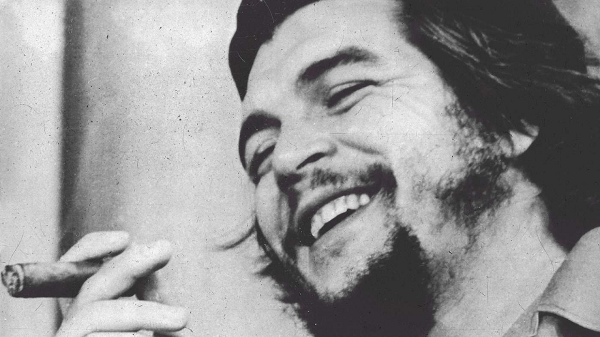 Hombre riendo y sosteniendo un cigarro, con barba y cabello largo, fondo desenfocado.
