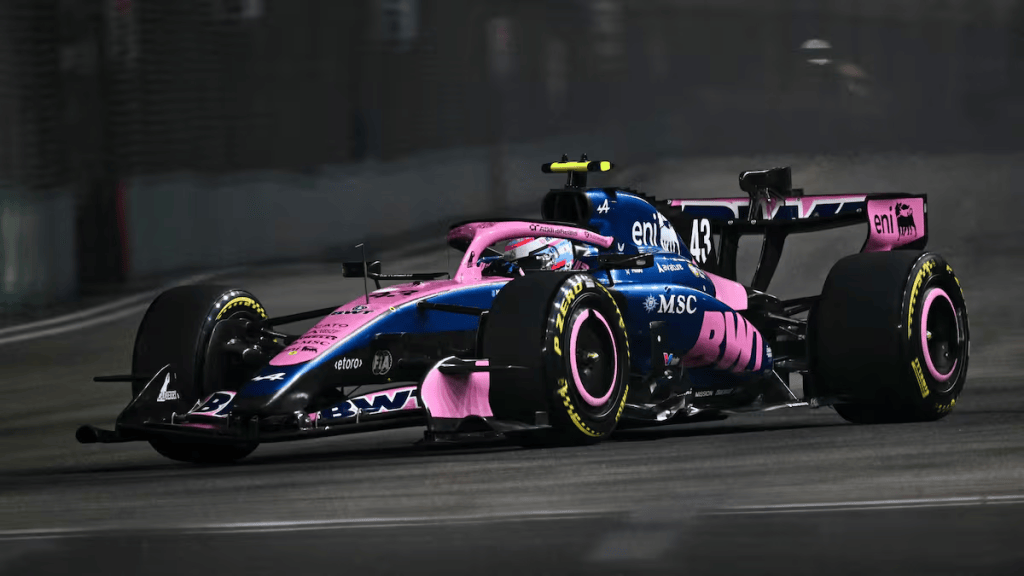 Coche de Fórmula 1 de color azul y rosa en una pista nocturna, con neumáticos amarillos destacados.
