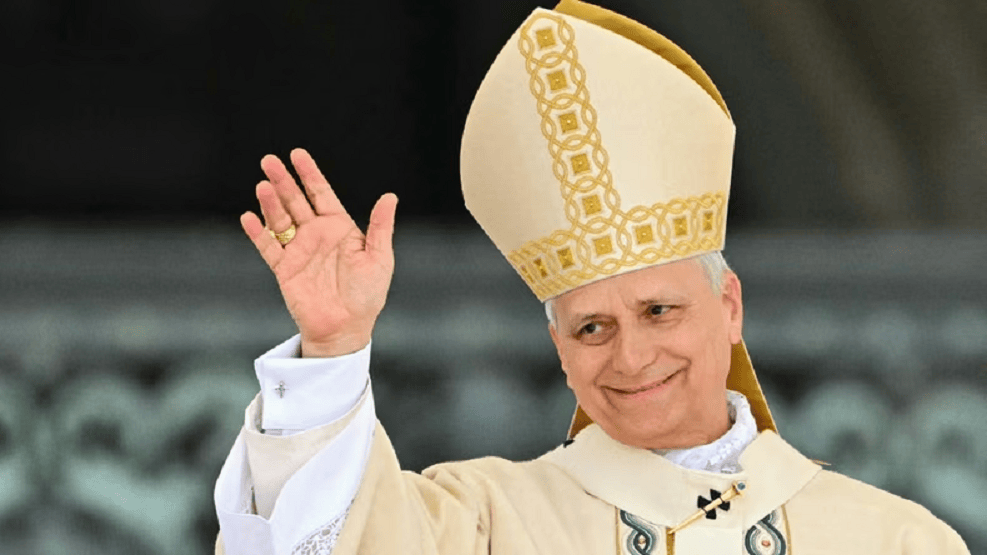 El Papa saludando con una mano, vistiendo una vestimenta papal y un gran sombrero blanco en un entorno al aire libre.