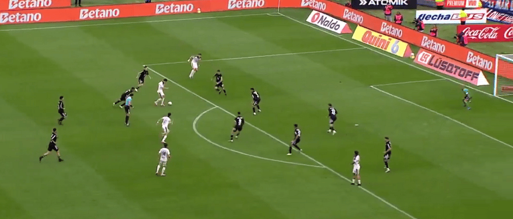 Juego de fútbol en el que un jugador en camiseta blanca está intentando avanzar hacia la portería mientras es marcado por varios defensores en camisetas negras.