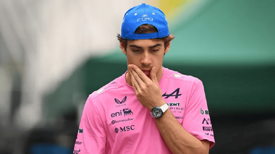 Joven piloto de Fórmula 1 vestido con una camiseta rosa y gorra azul, mostrando una expresión pensativa mientras se toca la boca.