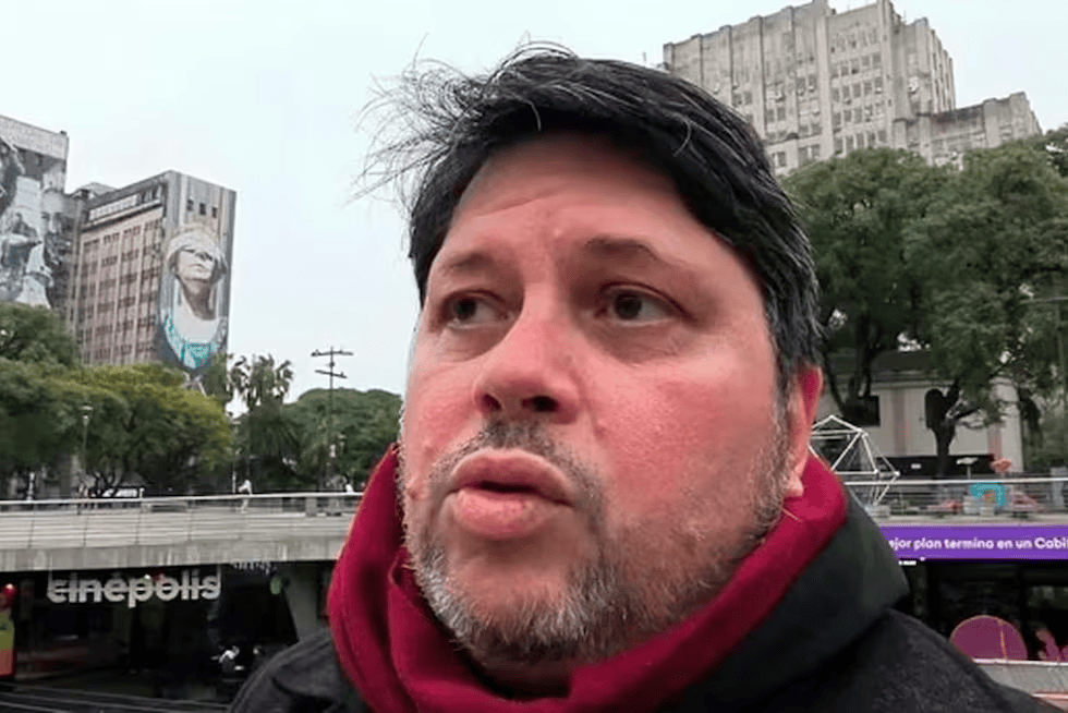 Un hombre con abrigo oscuro y bufanda roja habla sobre un fondo urbano con murales y edificios.