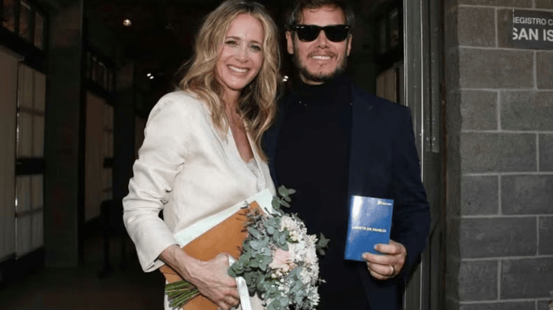 Pareja sonriente posando juntas, la mujer lleva un ramo de flores y un vestido claro, mientras el hombre viste una chaqueta oscura y sostiene un documento.