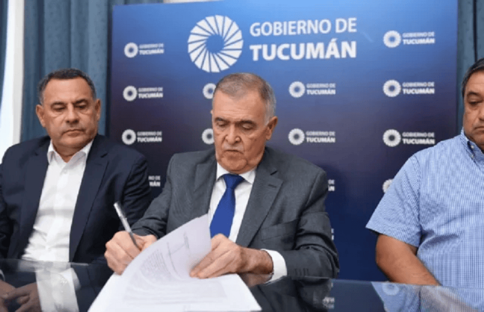 Un funcionario del Gobierno de Tucumán firma documentos mientras dos colegas lo observan, en un ambiente formal.