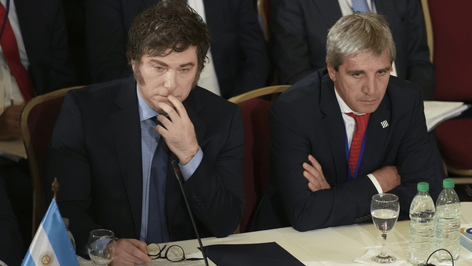 Dos hombres sentados en una mesa durante una reunión, ambos muestran expresiones reflexivas. Uno tiene el cabello rizado y viste un traje oscuro, mientras que el otro tiene cabello corto y rubio, vestido con un traje azul claro.