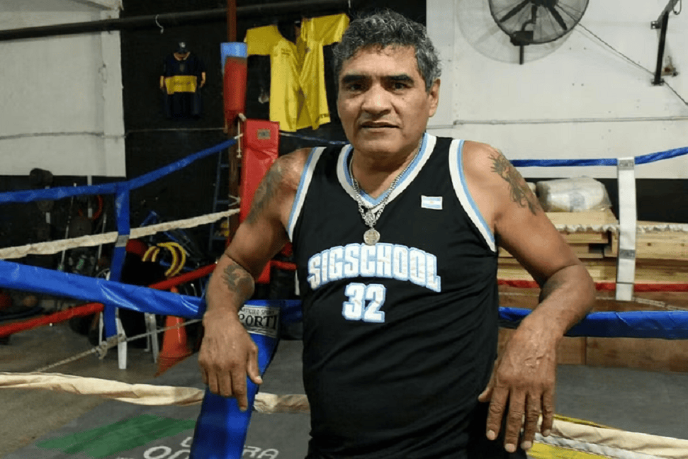 Hombre de pie en un gimnasio de boxeo, vistiendo una camiseta sin mangas negra con el número 32 y el texto 'SIGSCHOOL'.