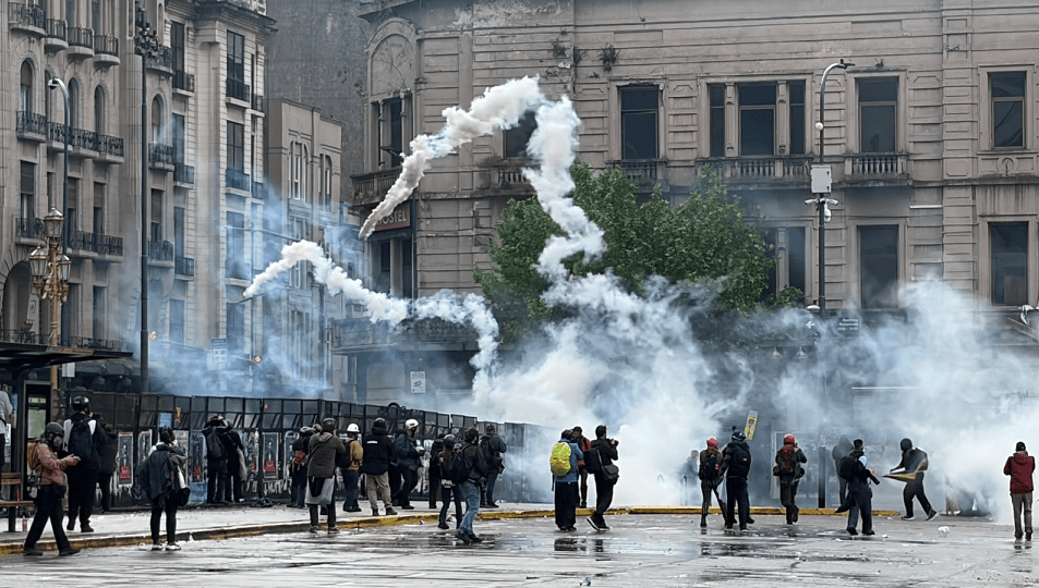 Manifestantes en una plaza rodeada de edificios, con humo de gas lacrimógeno y barricadas en un enfrentamiento con la policía.
