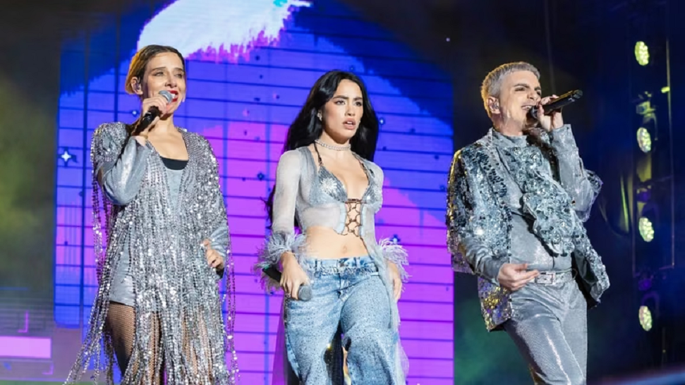 Tres artistas en un escenario, vestidos con trajes brillantes y cantando. El fondo muestra luces y una pantalla colorida.