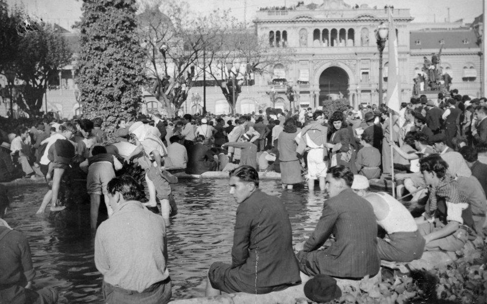 Una multitud de personas disfrutando en un parque alrededor de una fuente, algunos se mojan los pies en el agua, mientras otros observan y socializan en un día soleado.