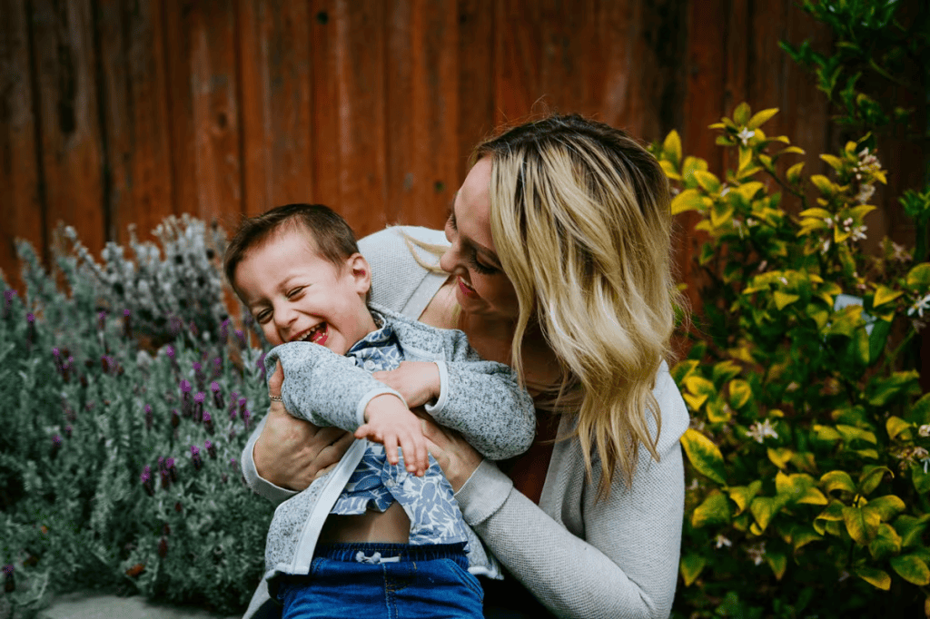Una madre sonriente sostiene a su hijo pequeño que ríe, rodeados de flores y un fondo de madera.