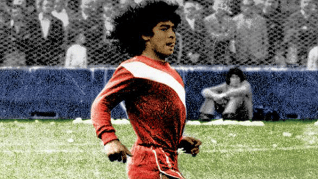 Jugador de fútbol corriendo en el campo, vistiendo una camiseta roja con una franja blanca.
