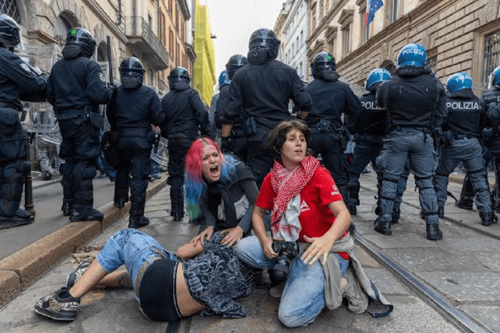 Manifestación con presencia de policías antidisturbios, dos mujeres en primer plano, una gritando y la otra agachada, con un ambiente tenso en la calle.