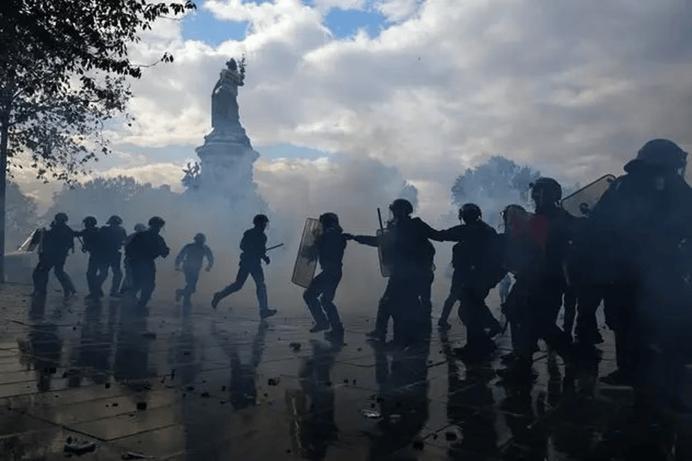 Protesta en una calle con humo y fuerza policial, personas corriendo y un monumento al fondo.