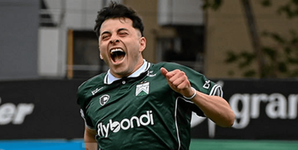 Jugador de fútbol celebrando efusivamente en un campo de juego, vistiendo una camiseta verde con el logo de un equipo.