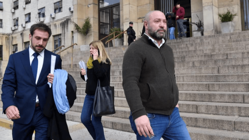 Tres personas saliendo de un edificio, con un hombre vestido de traje y dos personas más, una de ellas hablando por teléfono y la otra llevando documentos.