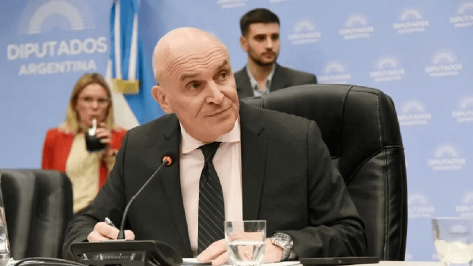 Un hombre de negocios sentado en una mesa, firmando documentos durante una reunión en un entorno gubernamental, con una mujer detrás sosteniendo una bebida y un hombre al fondo.