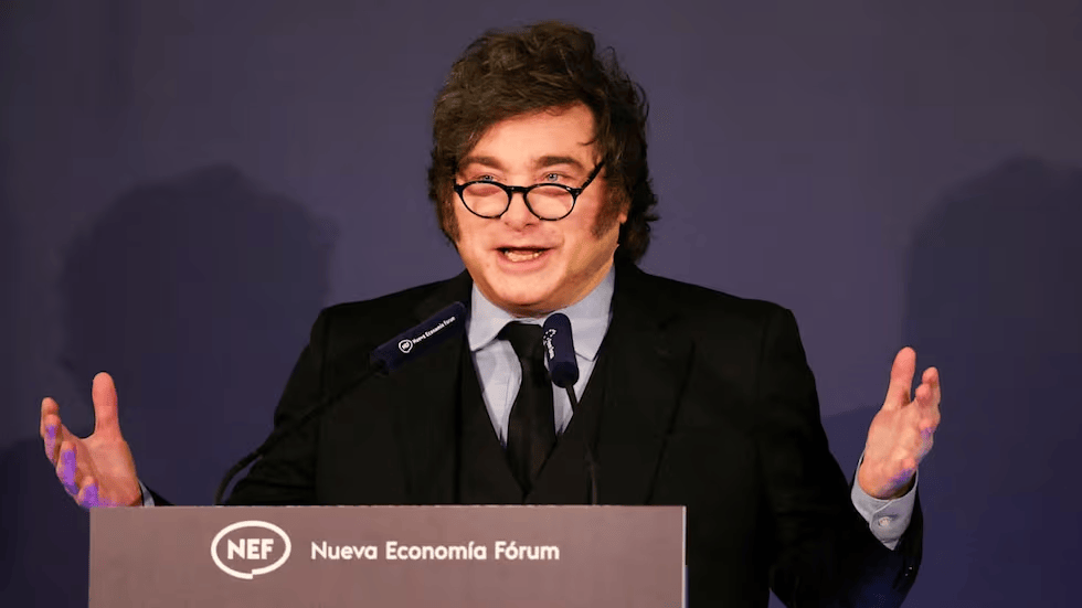 Hombre con cabello rizado y gafas, vestido de traje, hablando en un podio durante un evento del Nueva Economía Fórum.