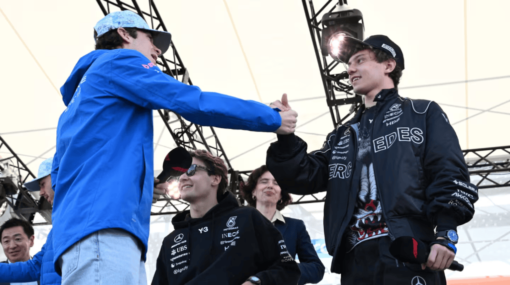 Dos jóvenes pilotos de automovilismo se dan la mano en un evento, mientras otros observan con sonrisas. Uno viste una chaqueta azul y el otro una chaqueta negra con logotipos.