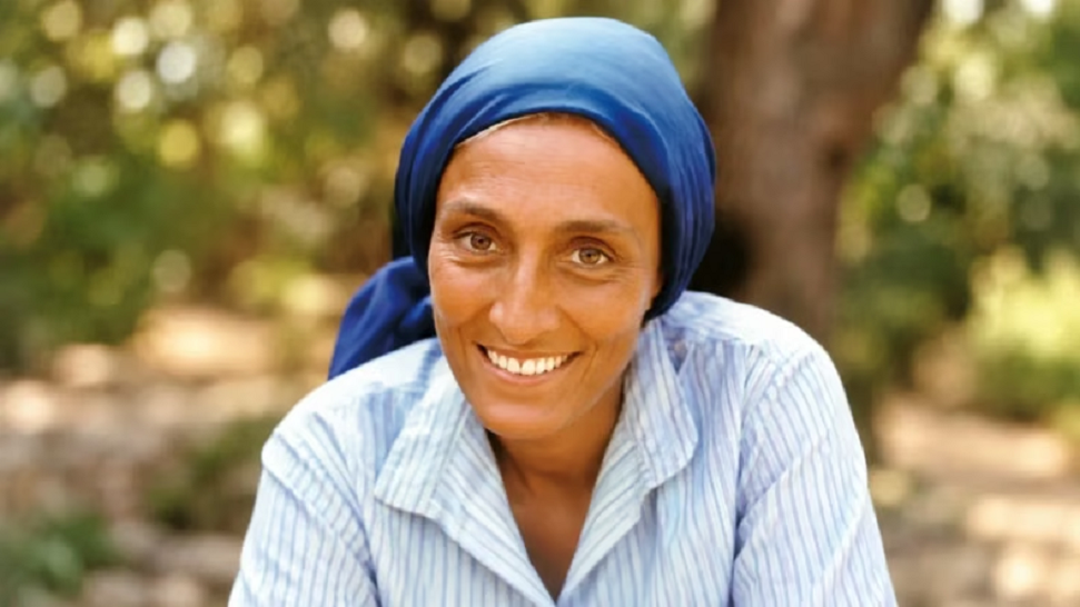 Mujer sonriendo, usando un pañuelo azul y una camisa a rayas, con un fondo natural difuminado.