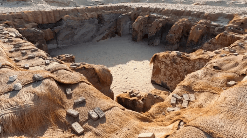 Vista de una excavación rectangular rodeada de arena y elementos de construcción antiguos, con hilos y bloques de material en la superficie.