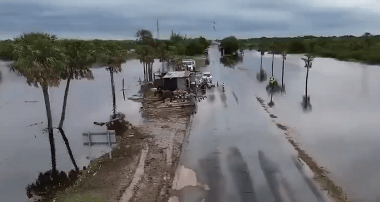 Inundación en una carretera, con árboles y estructuras sumergidos en agua, bajo un cielo nublado.