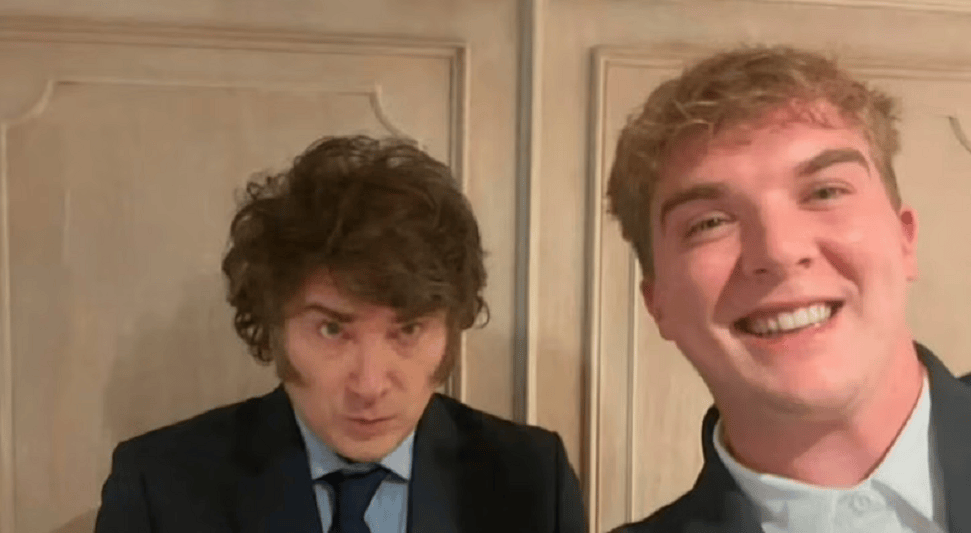 Dos hombres posando para una selfie, uno con cabello rizado y serio, y el otro sonriendo ampliamente, ambos en trajes formales.