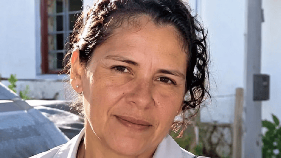 Primer plano de una mujer con cabello rizado, sonriendo ligeramente, en un entorno exterior con una casa de fondo.
