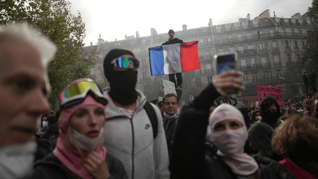 Manifestación con personas enmascaradas y un hombre de pie sosteniendo una bandera francesa en un ambiente nublado.