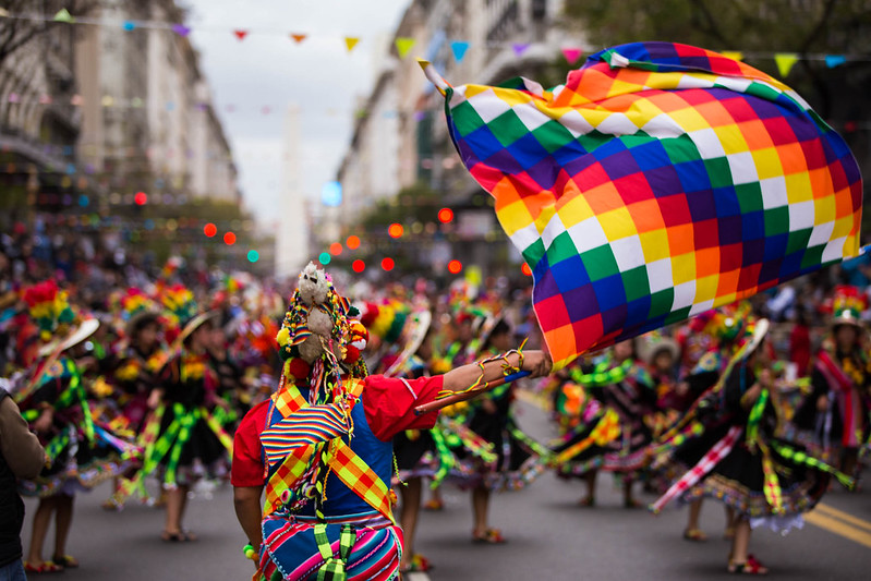 Grupo de bailarines en un desfile, vestidos con trajes coloridos y sosteniendo banderas multicolores, en una calle bulliciosa.